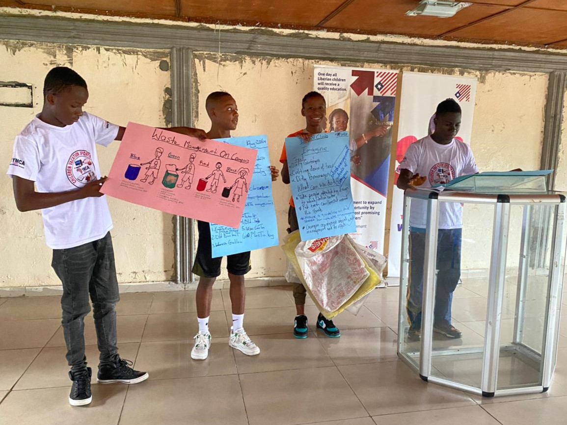 Four Black boys hold up posters about waste management at the front of a room